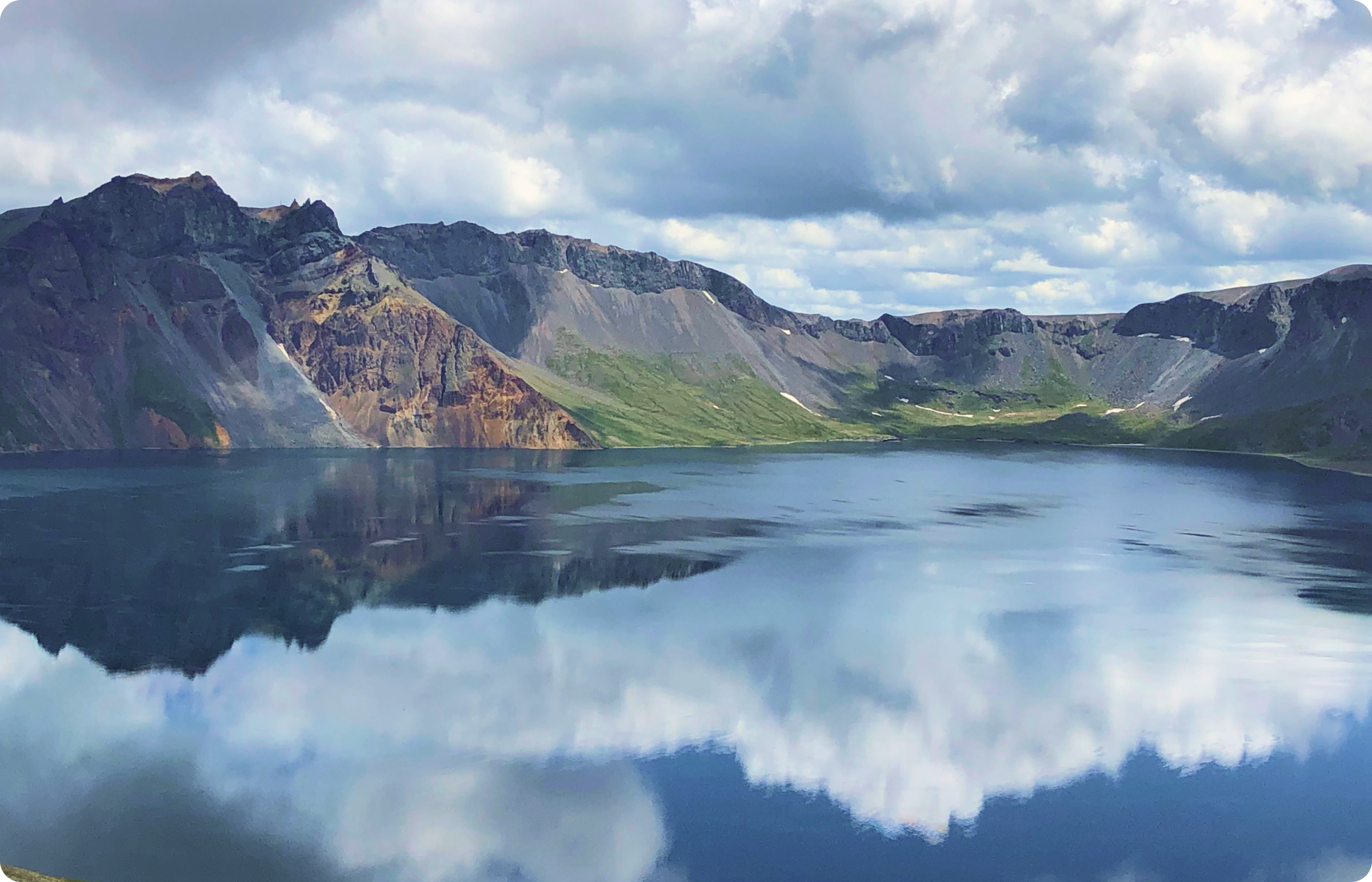 雲が反映する穏やかな湖の水面と、その背後にそびえる緑豊かな山並みの景色